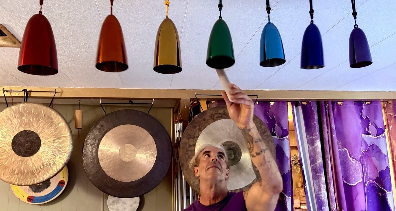 Vance sounding the row of rainbow chakra chimes above the table, gongs on the wall behind him