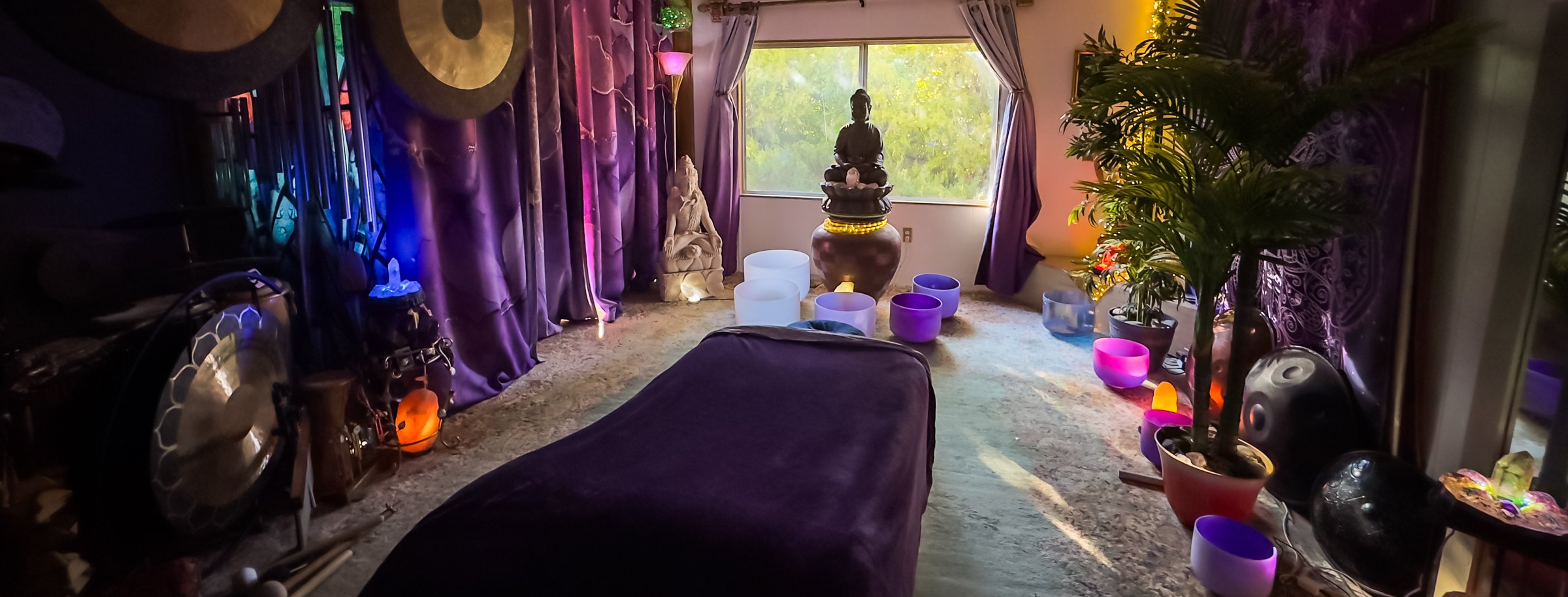The healing room at golden hour — gongs along the wall, the Buddha fountain in the window, the table dressed in purple